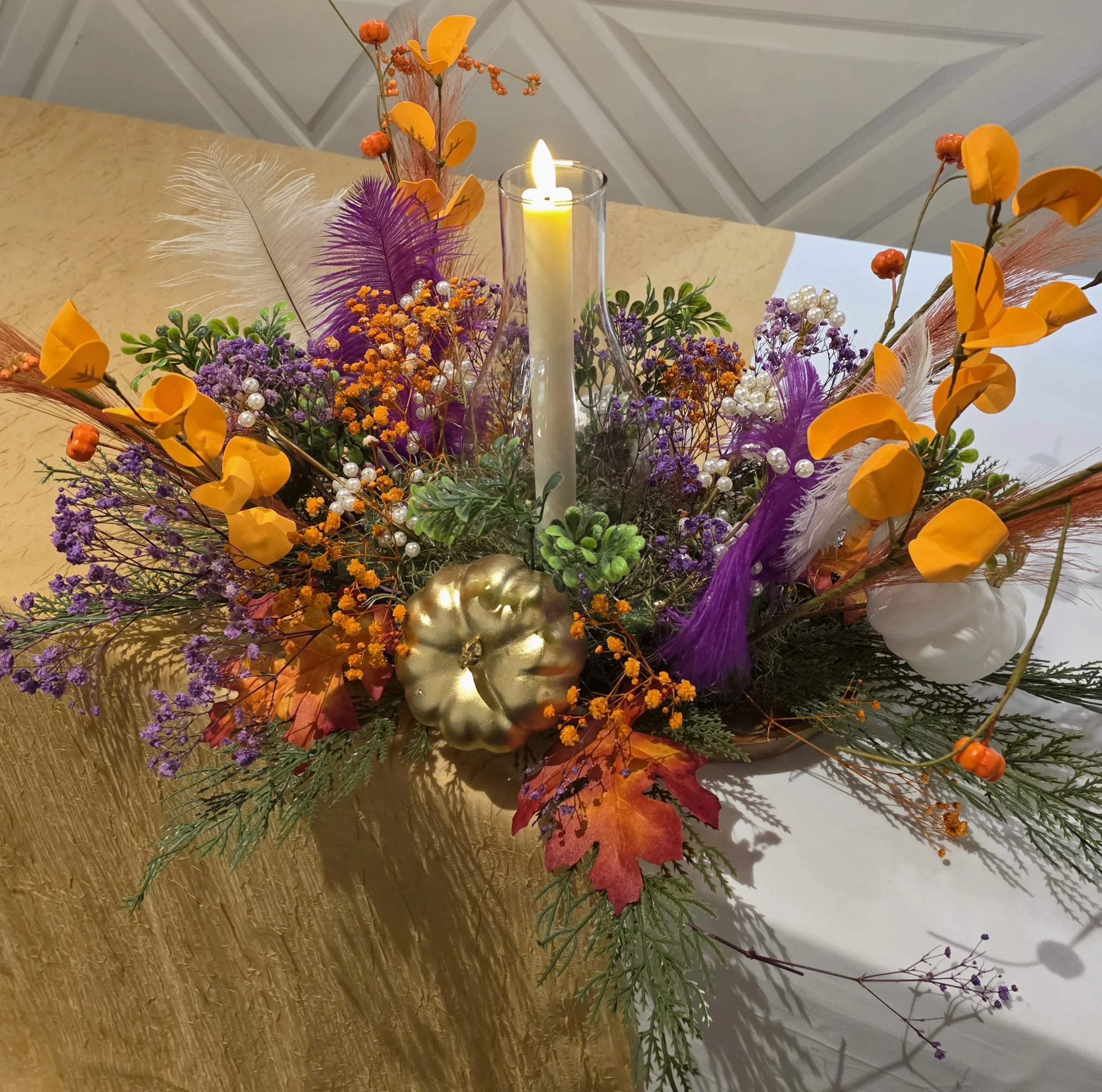 A floral arrangement with purple, orange, white, and green flowers and plants, centered around a white candle in a glass holder, with golden decorative pumpkins at the base, on a table with a beige tablecloth.