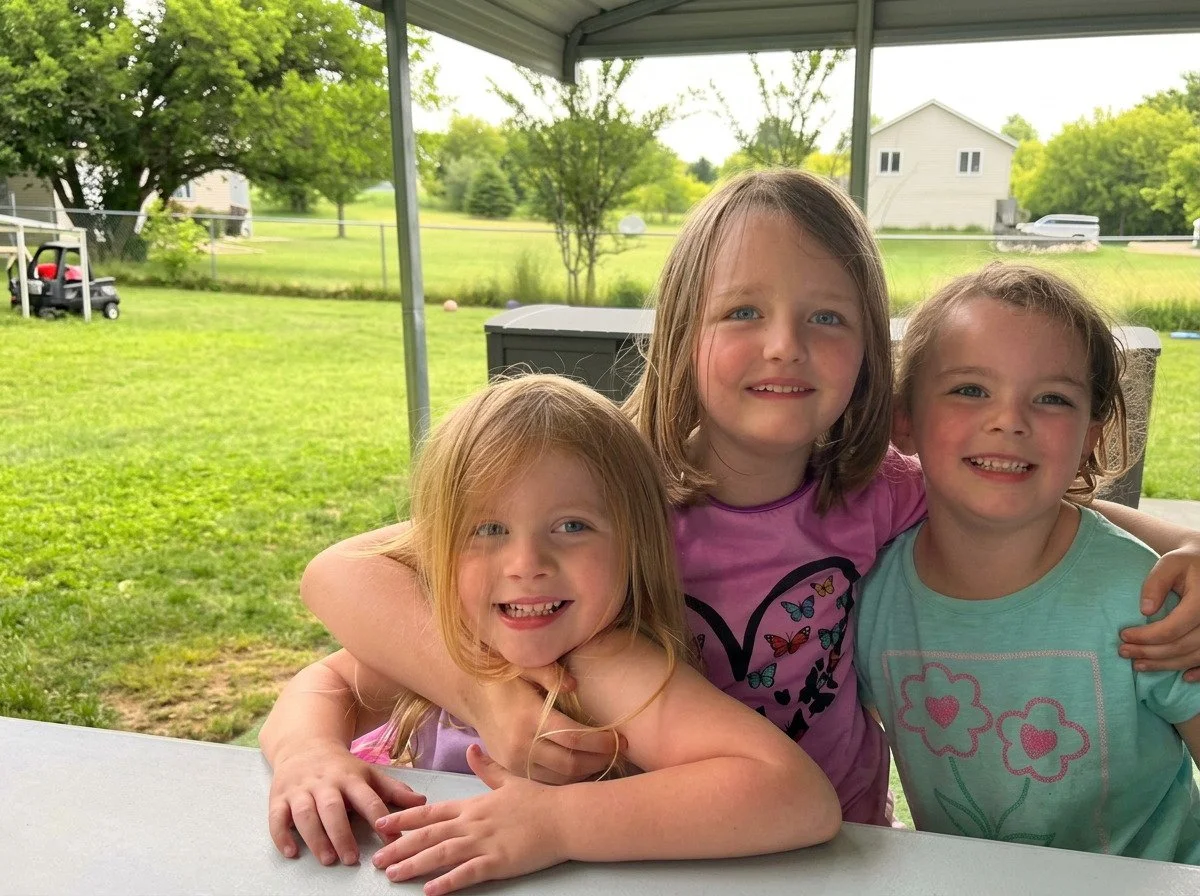Three children smiling together at a picnic table in the fenced backyard at Little Monkeys Child Care.