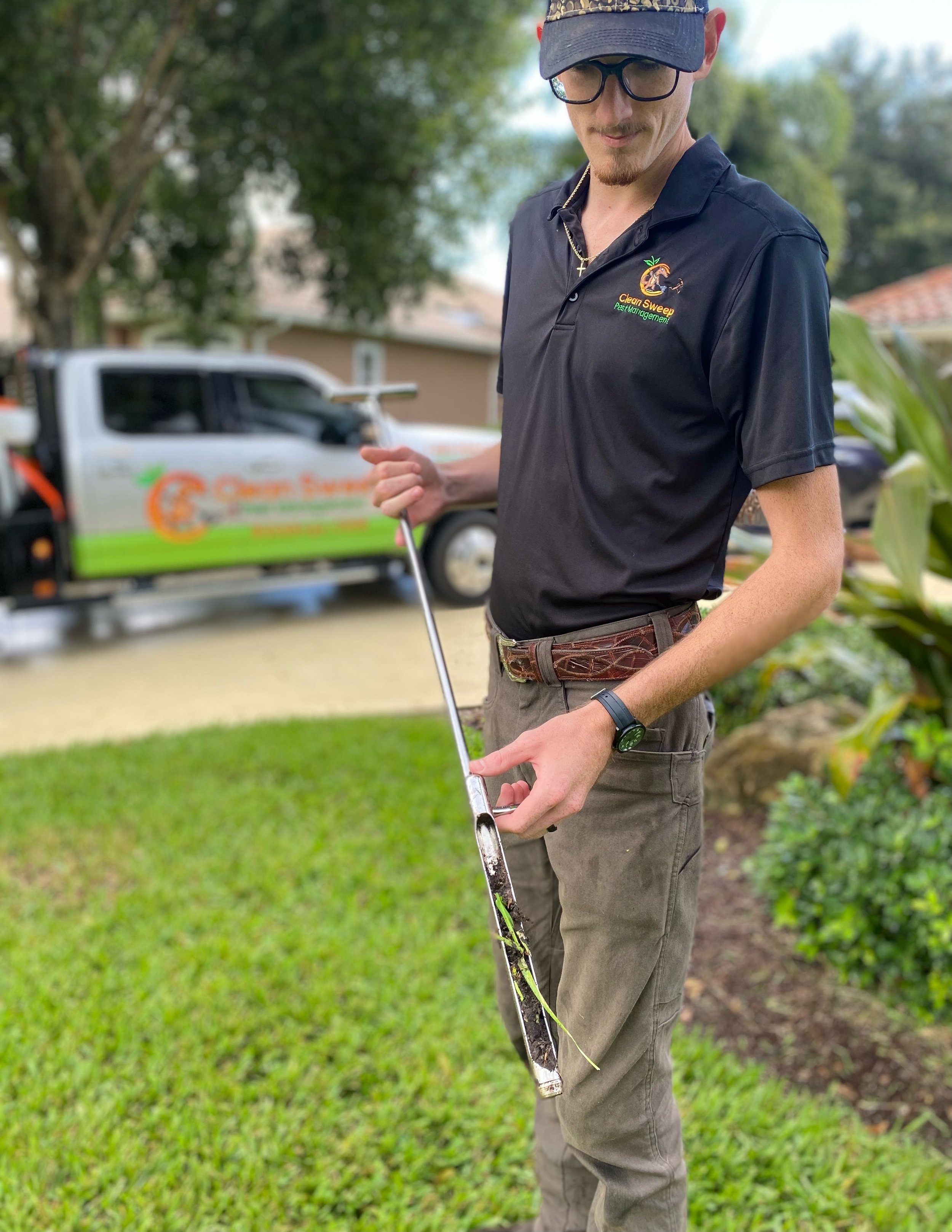 Man outdoors holding a measuring tape, wearing glasses, a black polo with a company logo, and a watch, with a truck and trees in the background.
