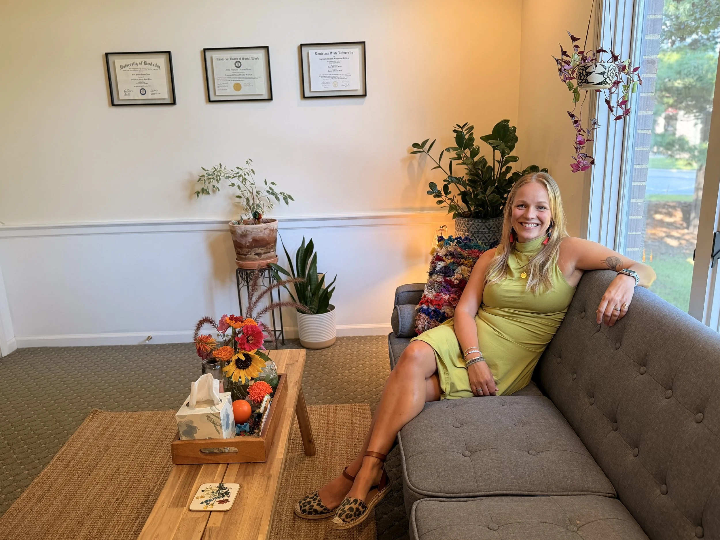 A woman with blonde hair smiling, sitting on a gray couch in a well-lit room with a large window. The room has several potted plants, framed certificates on the wall, a wooden coffee table with flowers and decorative items, and a colorful cushion behind her.