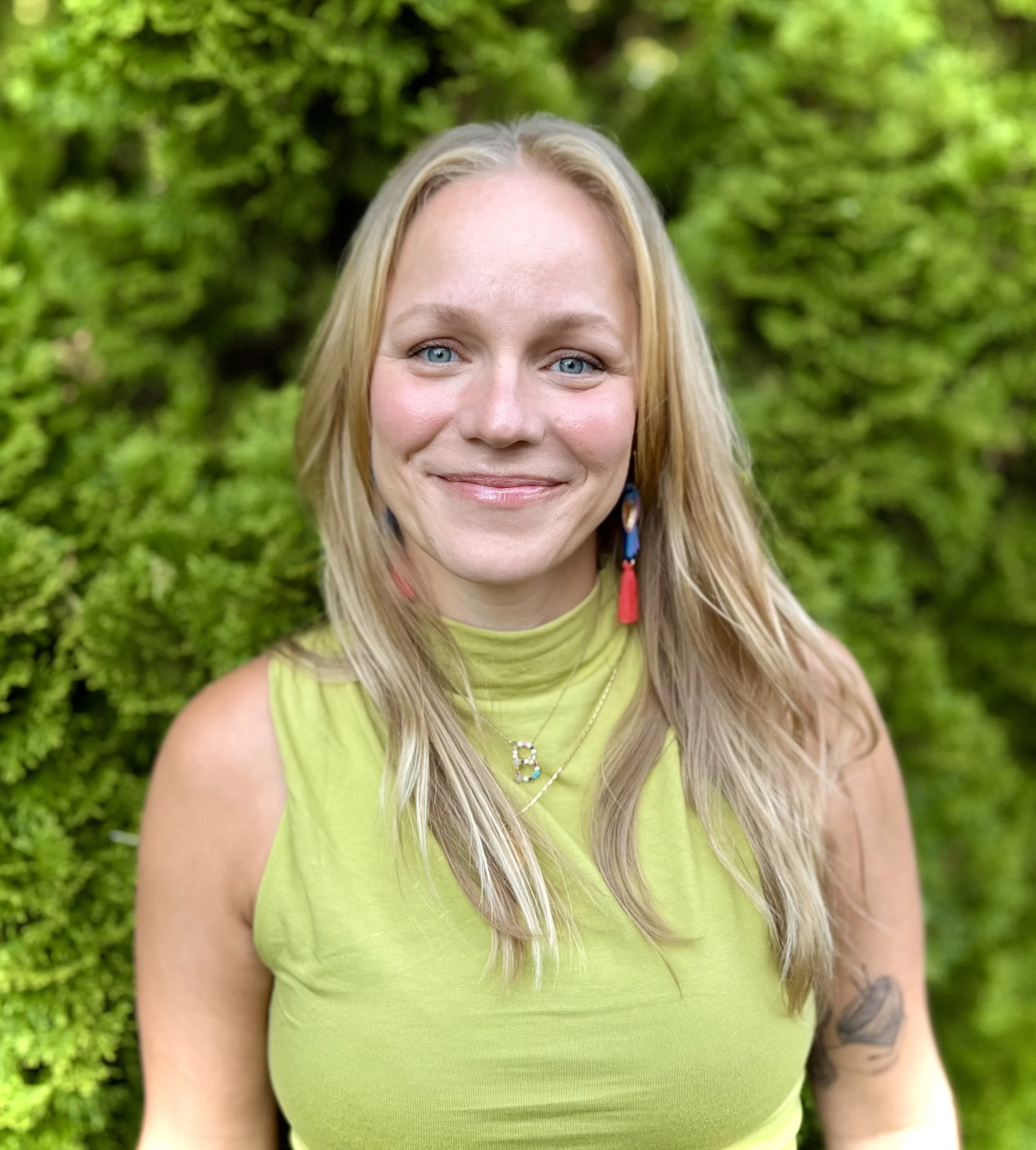 Close-up of a smiling woman with long blonde hair and blue eyes, wearing a light green sleeveless top, standing outdoors with a blurred green foliage background.