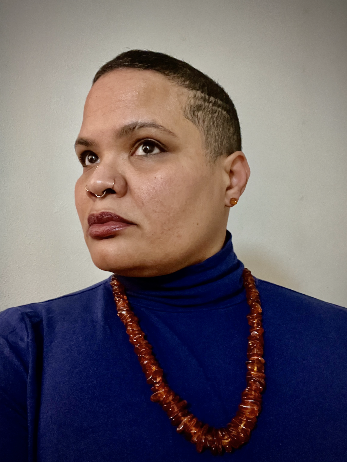 Person with short dark hair wearing a blue turtleneck, amber-colored beaded necklace, and earrings, looking to the left with a neutral expression against a light gray background.