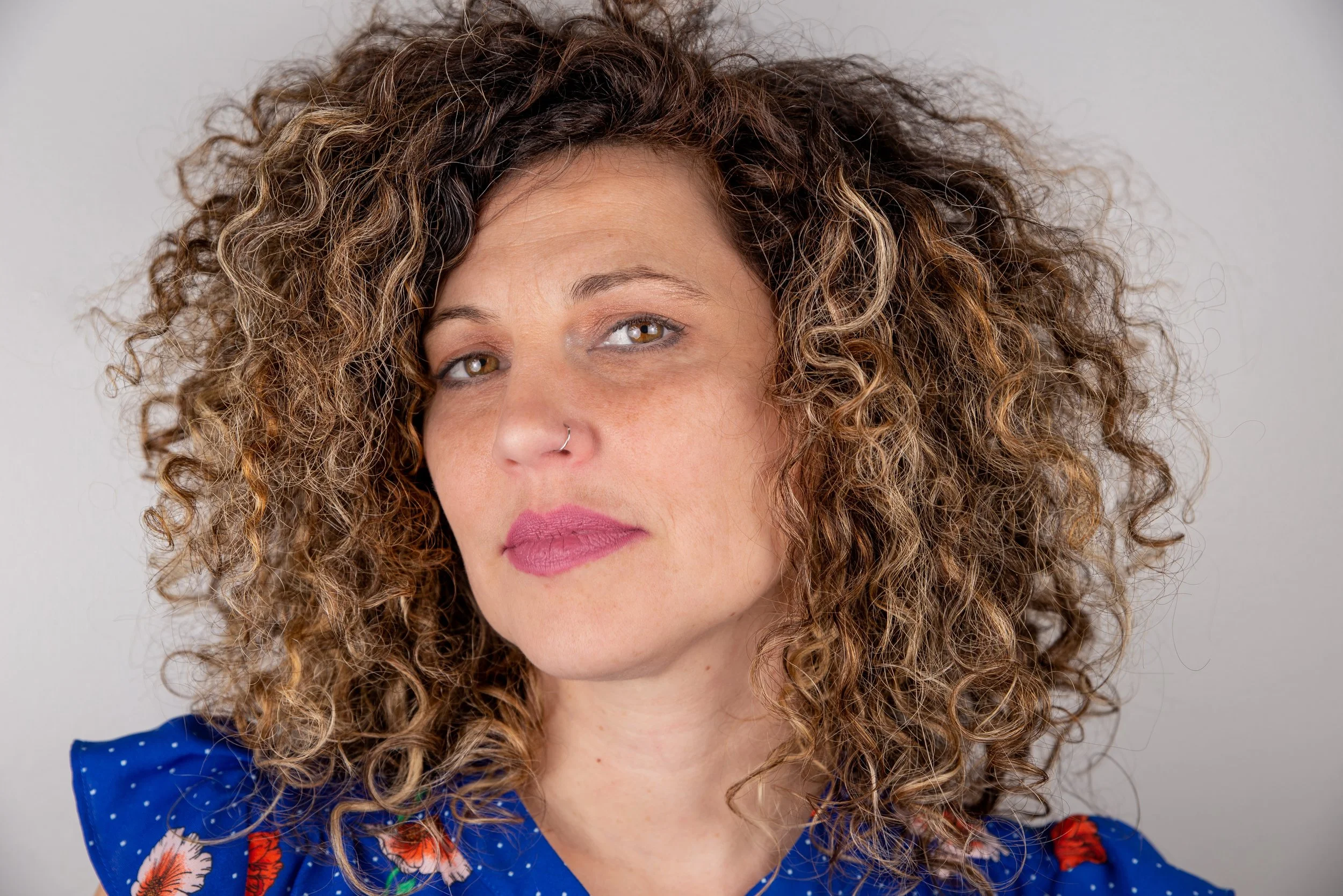 Close-up of a woman with curly brown hair and light brown eyes, wearing pink lipstick and a nose ring. She is dressed in a blue top with a floral pattern.