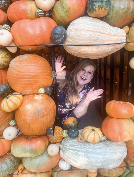 A woman smiling and waving from behind a colorful arrangement of pumpkins and gourds of various sizes and colors.