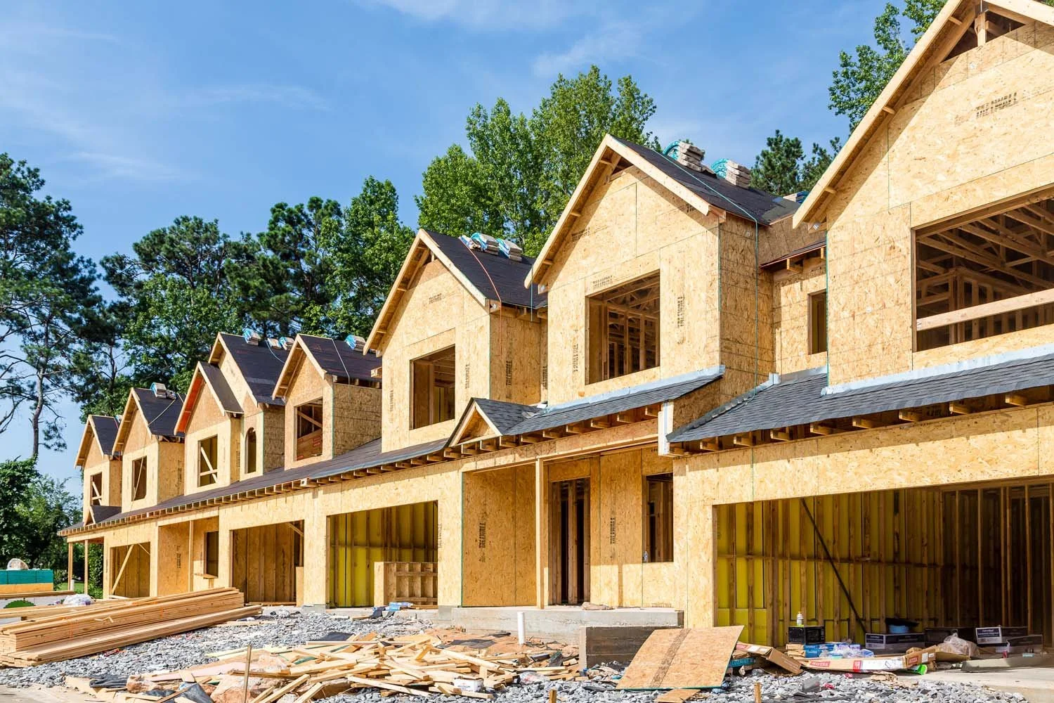 Row of new townhouses under construction with exposed wooden frames and roofing in progress, surrounded by construction debris, trees, and a clear blue sky.