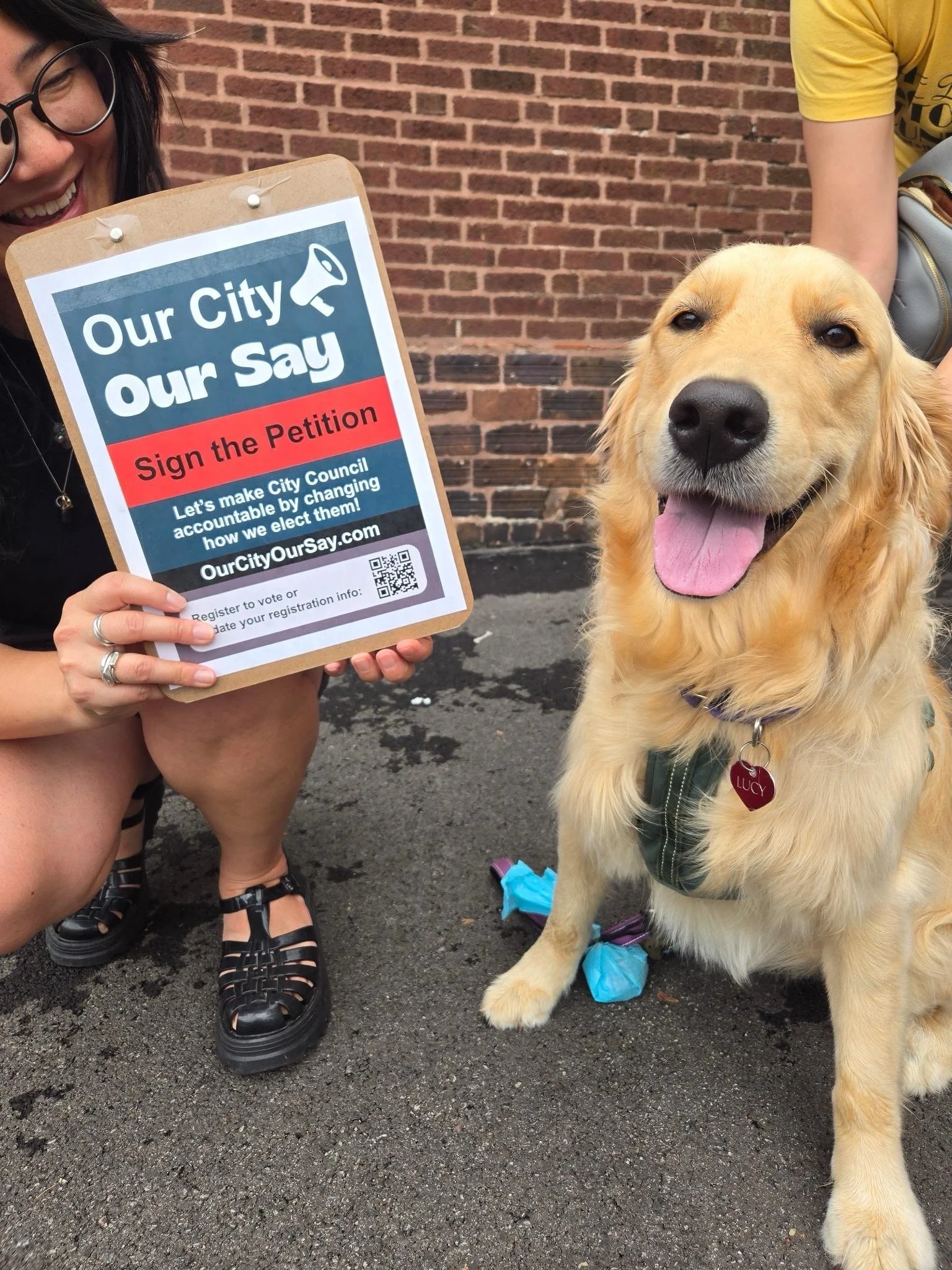 A woman with glasses smiling holding a sign that reads 'Our City Our Say Sign the Petition' standing next to a golden retriever with a purple collar, black harness, and a red heart-shaped tag with the name Lucy, against a brick wall background.
