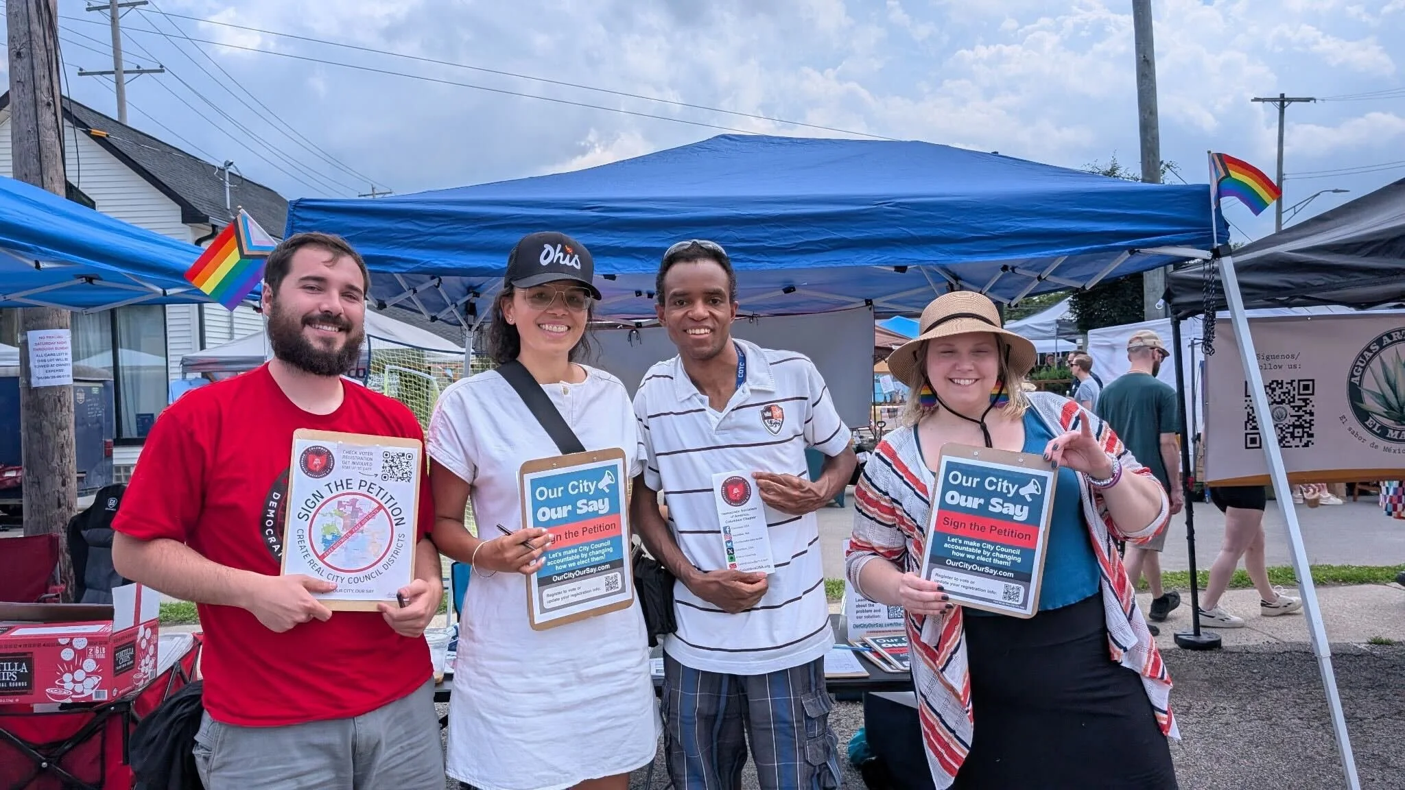 Four people standing together at an outdoor event holding signs supporting city council elections, under a blue canopy tent decorated with rainbow flags, with other tents and people in the background.