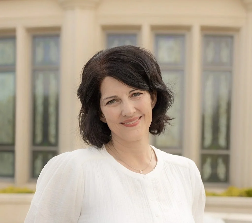 A woman with black hair and a white blouse standing outdoors in front of a building with large windows and columns.