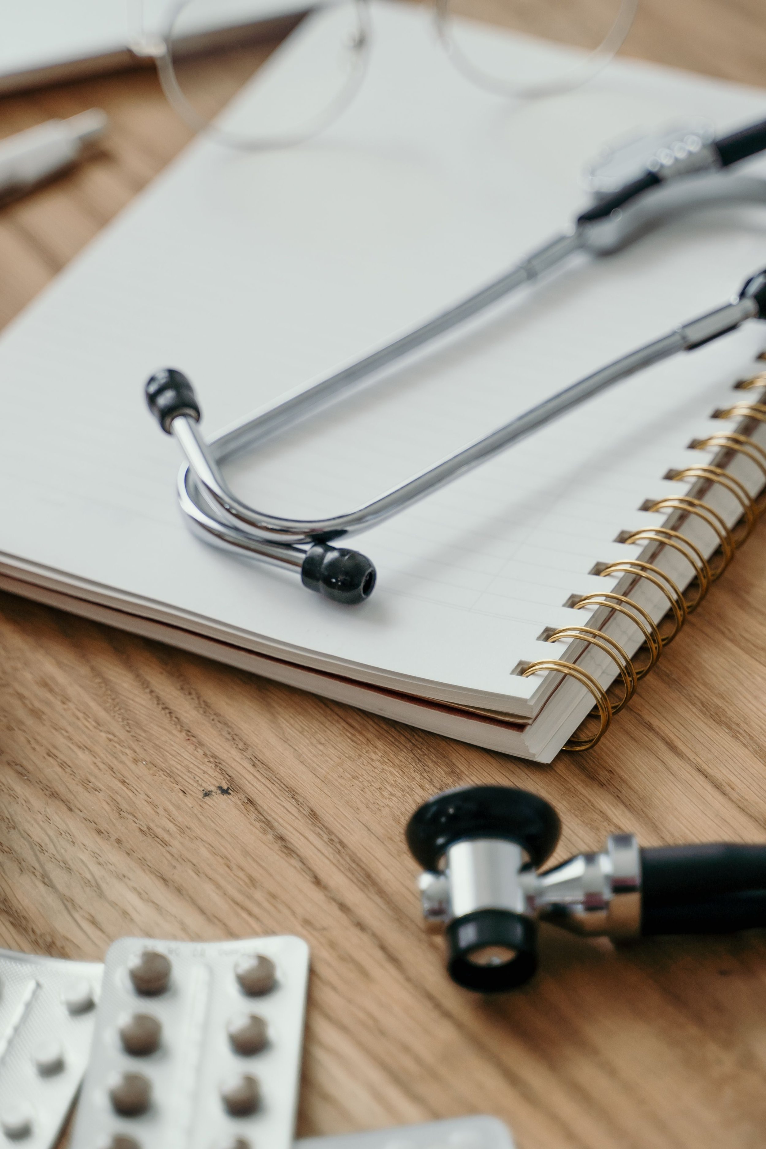 Close-up of a wooden desk with medication blister packs, a white notepad, metal medical tools, and eyeglasses.