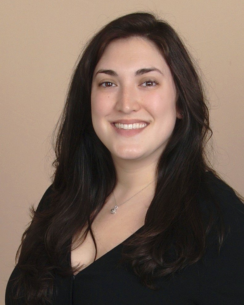 A young woman with long dark hair smiling, wearing a black top and a silver necklace with a small pendant, standing against a neutral background.