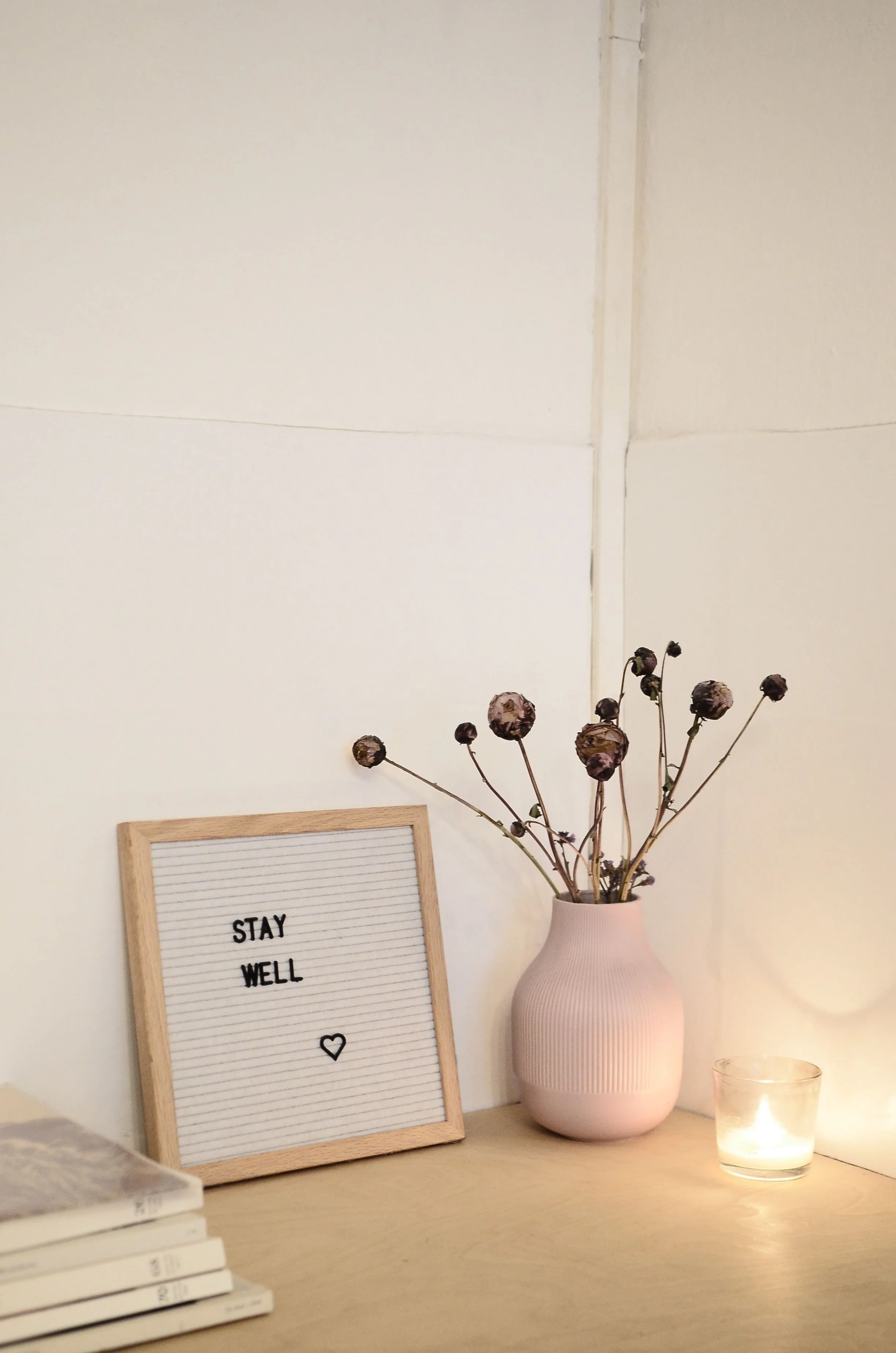 Decorative desk setup with a light wood framed letter board displaying the message 'STAY WELL'  with a small black heart, a pink ceramic vase with dried dark purple flowers, a small candle holder with a lit candle, and a partial view of a book stack.