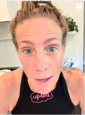 Close-up of a woman with red hair and blue eyes, wearing a black tank top, with a tattoo on her shoulder, indoors with a plant and a shelf in the background.