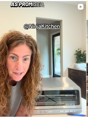 Woman with curly hair in a kitchen, near a toaster oven, with a plant on a cabinet and a door in the background.