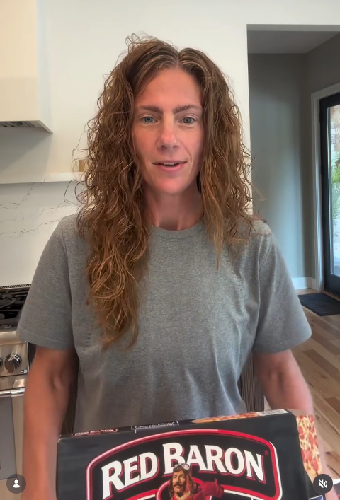 Woman with curly red hair wearing a gray t-shirt, standing in a kitchen, holding a box of Red Baron pizza.