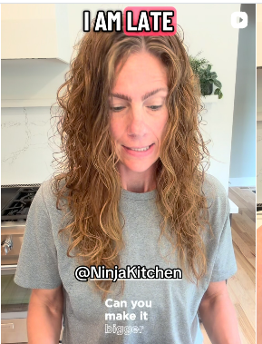 Woman with curly hair in a kitchen, wearing a gray t-shirt, looking down, with text overlay saying 'I am late' and '@NinjaKitchen'.