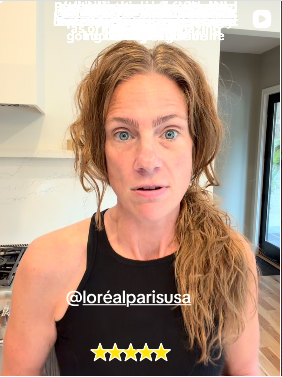 A woman with wavy, light brown hair and blue eyes wearing a black tank top, standing in a modern kitchen with a white wall and door in the background.