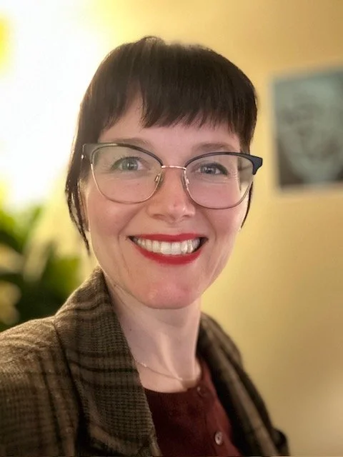 A woman with short dark hair, glasses, and red lipstick smiling for a selfie. She is wearing a brown plaid blazer over a maroon shirt.