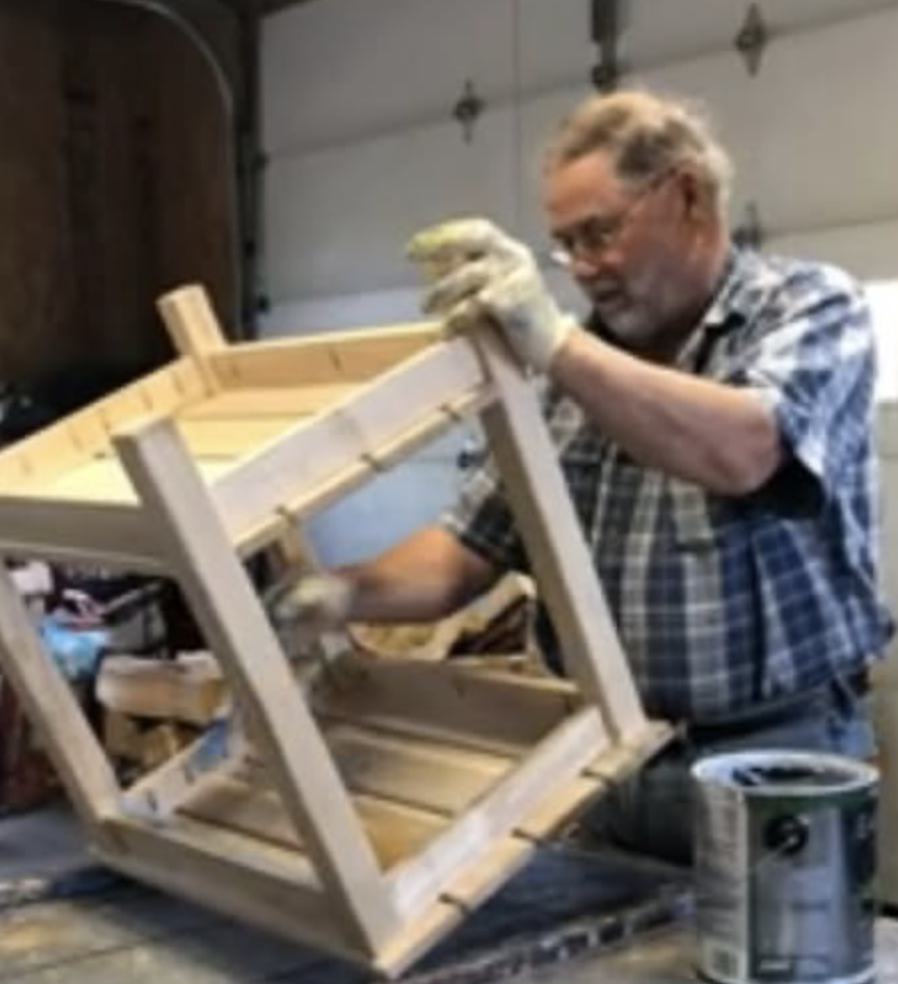 An older man wearing glasses, a plaid shirt, and work gloves is painting a wooden chair in a garage workshop.