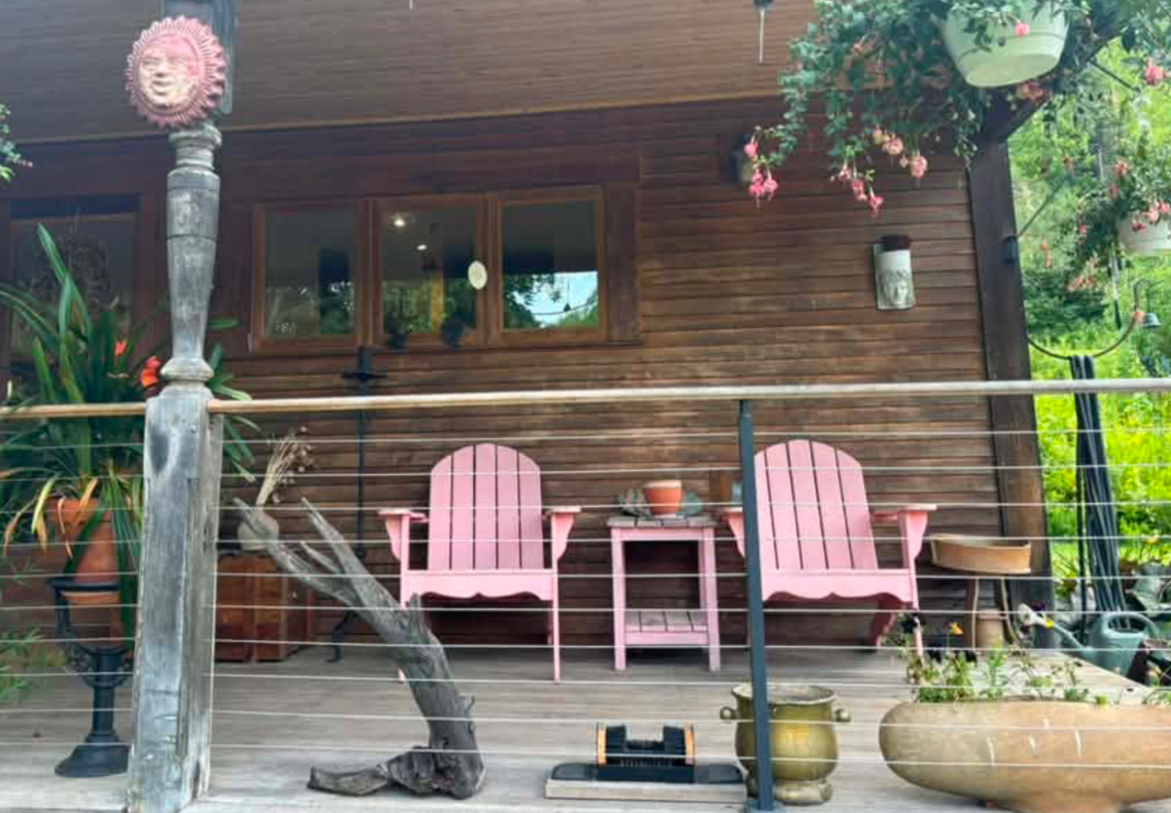 A front porch of a house with wooden exterior walls, pink Adirondack chairs, potted plants, and hanging flower baskets. There is a tiki torch in the foreground and decorative items hanging on the wall. The porch is enclosed with a wire fence and surr