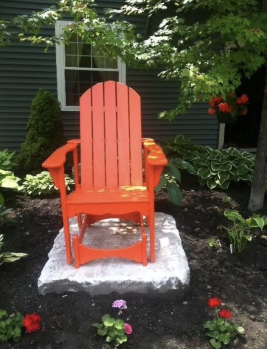 Orange Adirondack chair on a stone platform in a garden with green plants and flowers, in front of a house with dark siding and a window.