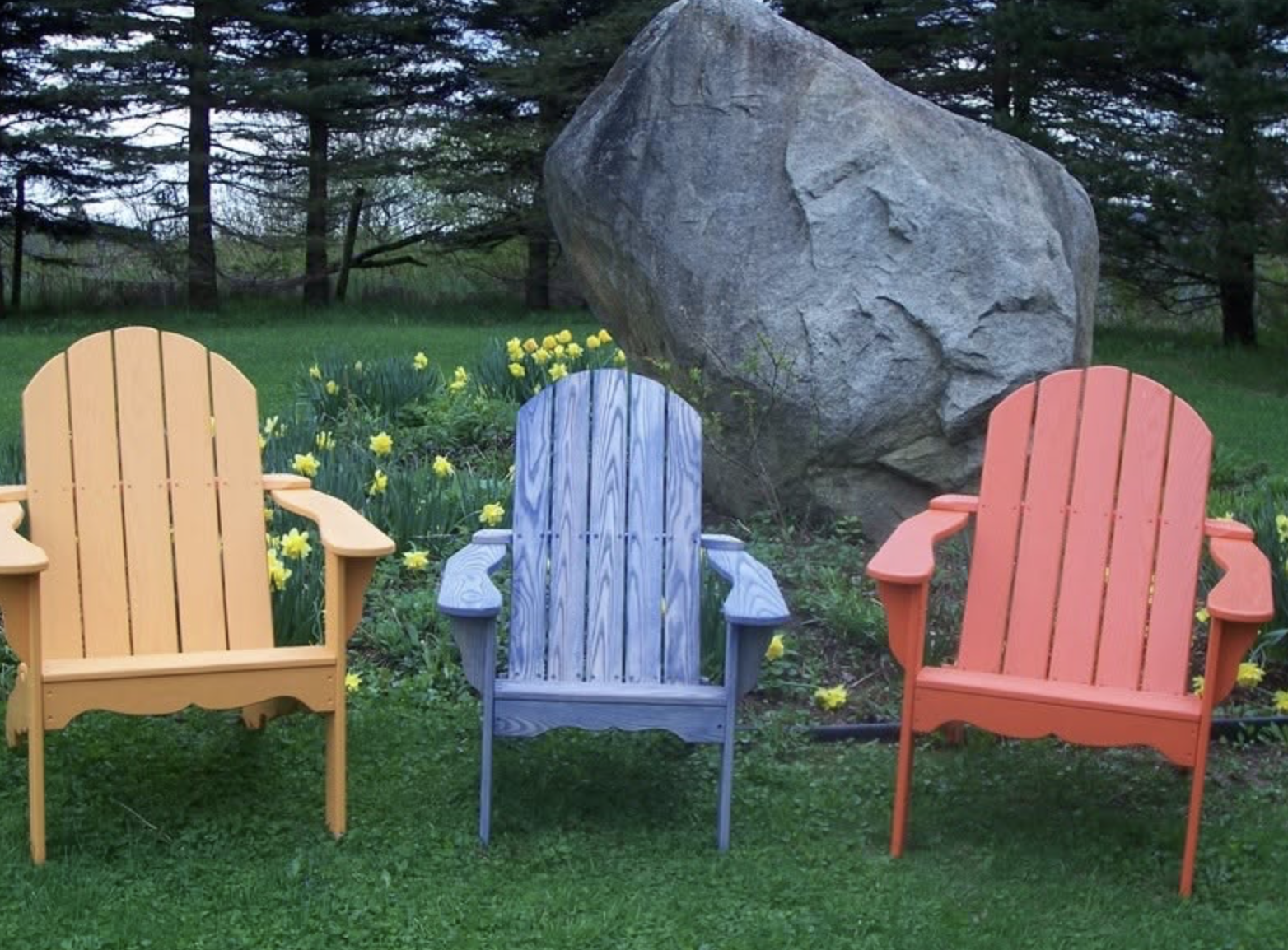 Three colorful Adirondack chairs—yellow, gray, and orange—placed outdoors on green grass with yellow daffodils and a large boulder in the background.