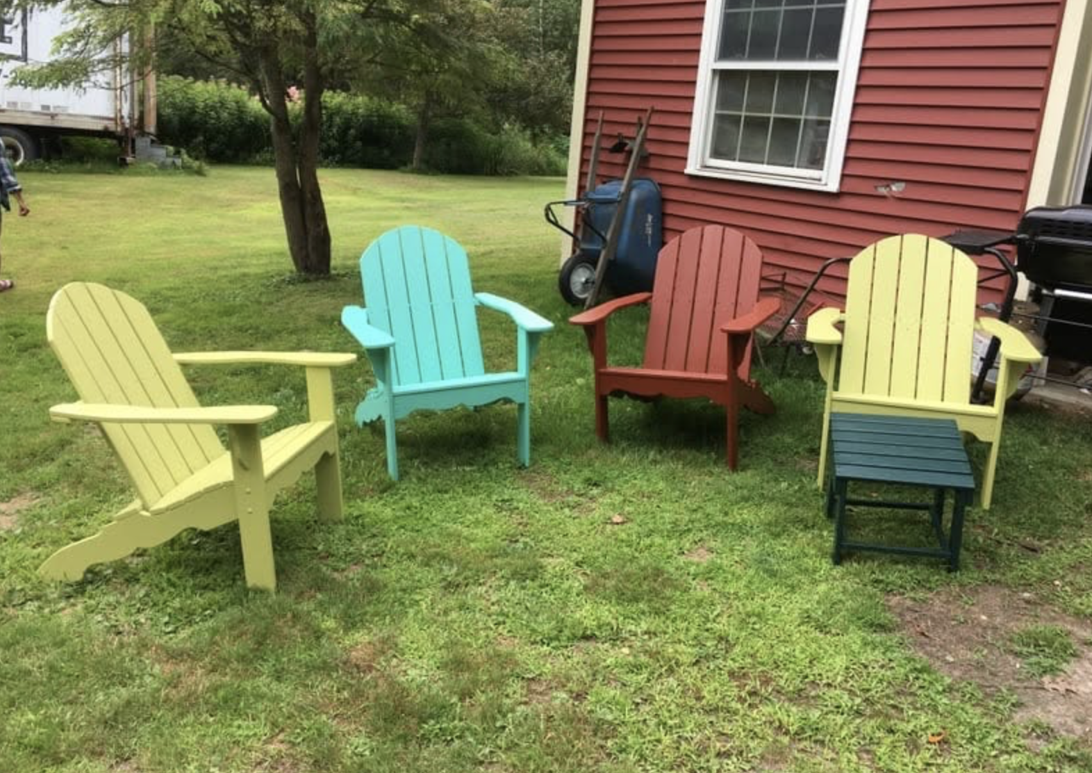 Colorful outdoor Adirondack chairs in a backyard with green grass, a tree, and a red house wall, accompanied by a wheelbarrow and a grill.