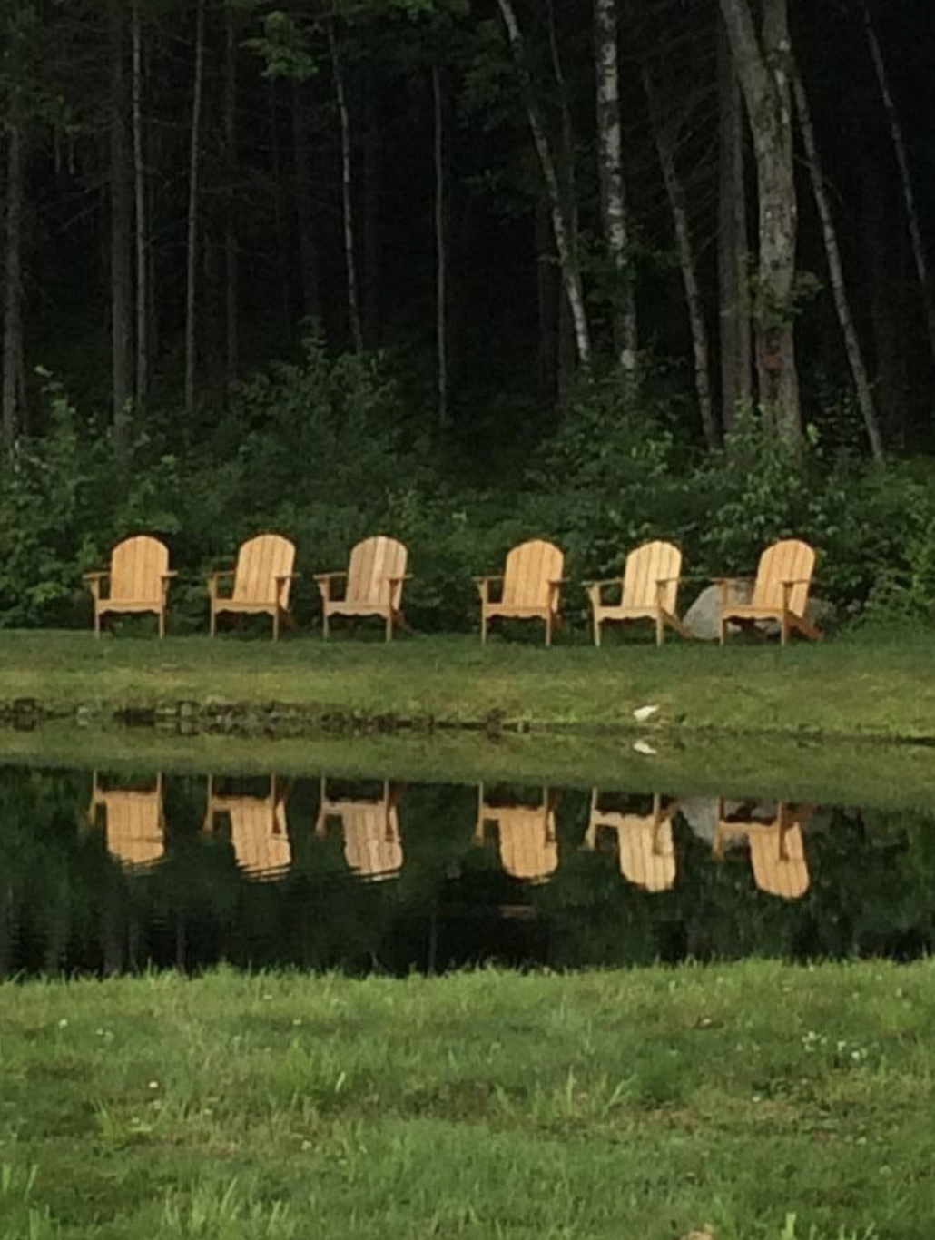 Six wooden Adirondack chairs lined up by a lake, with their reflections visible in the water, set against a background of trees and greenery at dusk or evening.