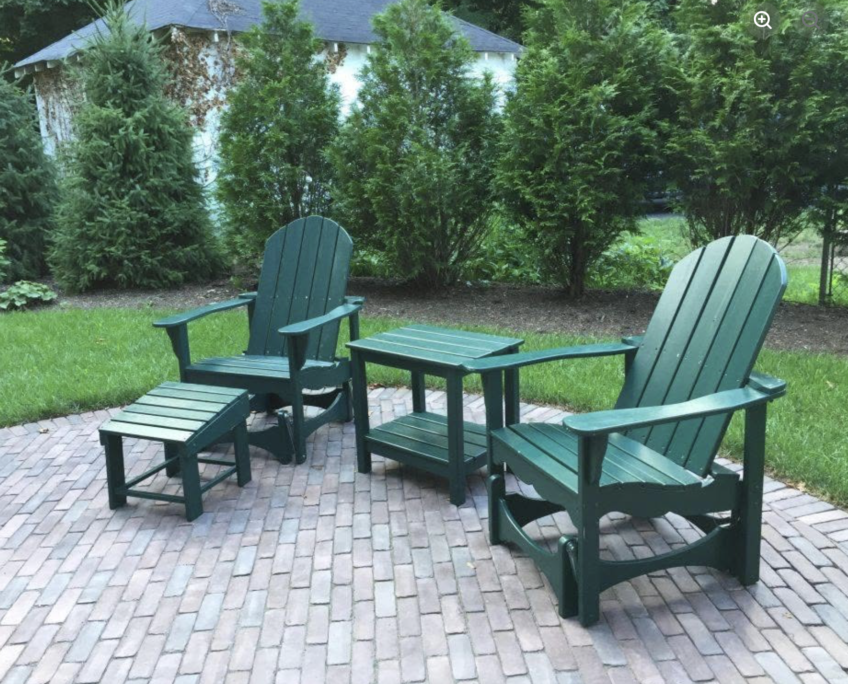 Green outdoor furniture including two Adirondack chairs, a matching footstool, and a small table on a brick patio, surrounded by lush green bushes and trees.