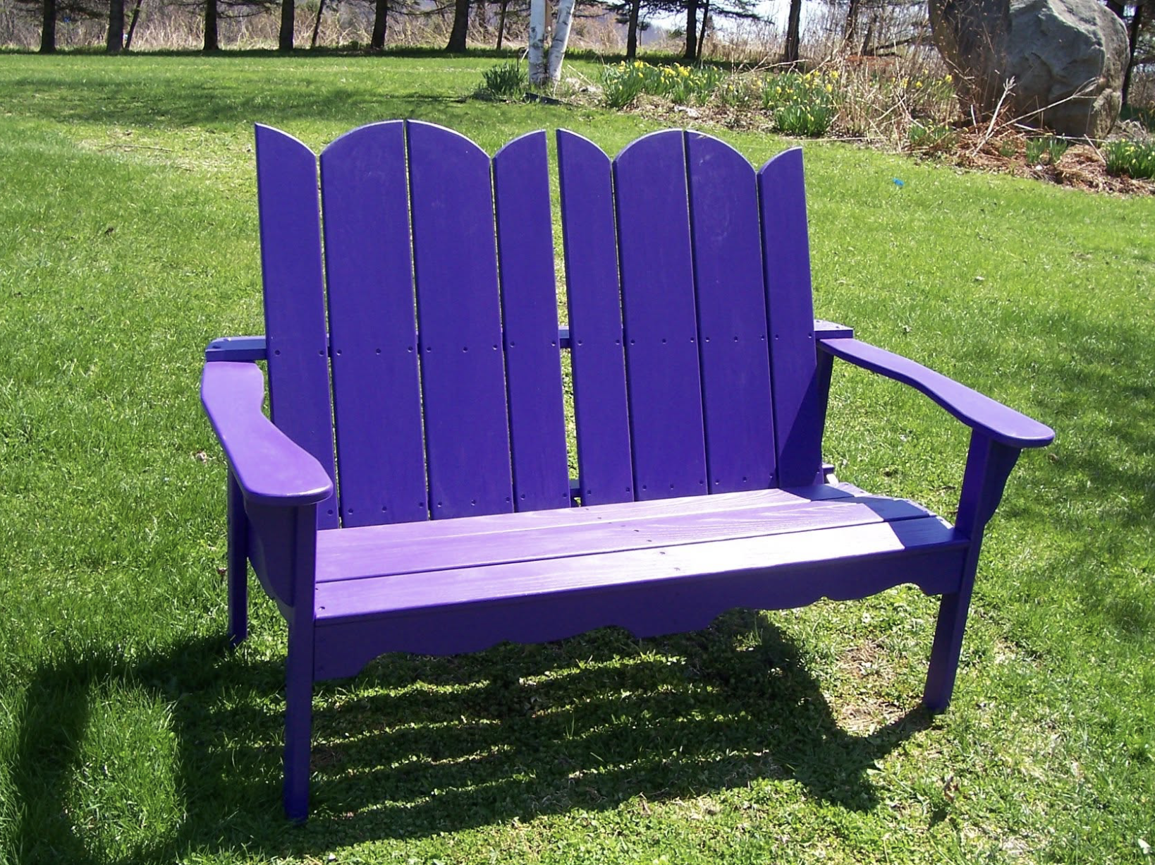 A purple wooden bench on a grassy lawn with trees and rocks in the background.