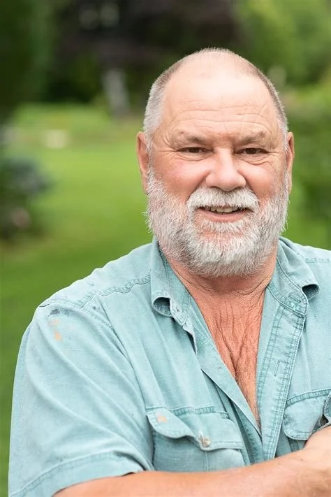 A smiling elderly man with a white beard and mustache, wearing a light blue button-up shirt, outdoors with green trees and grass in the background.