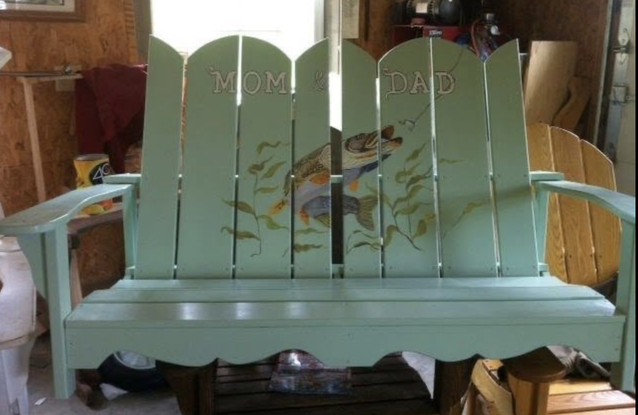 A painted wooden Adirondack loveseat with a handprinted trout angling towards a fly fishing lure,  and the words 'Mom & Dad' painted on the backrest, situated in a workshop.