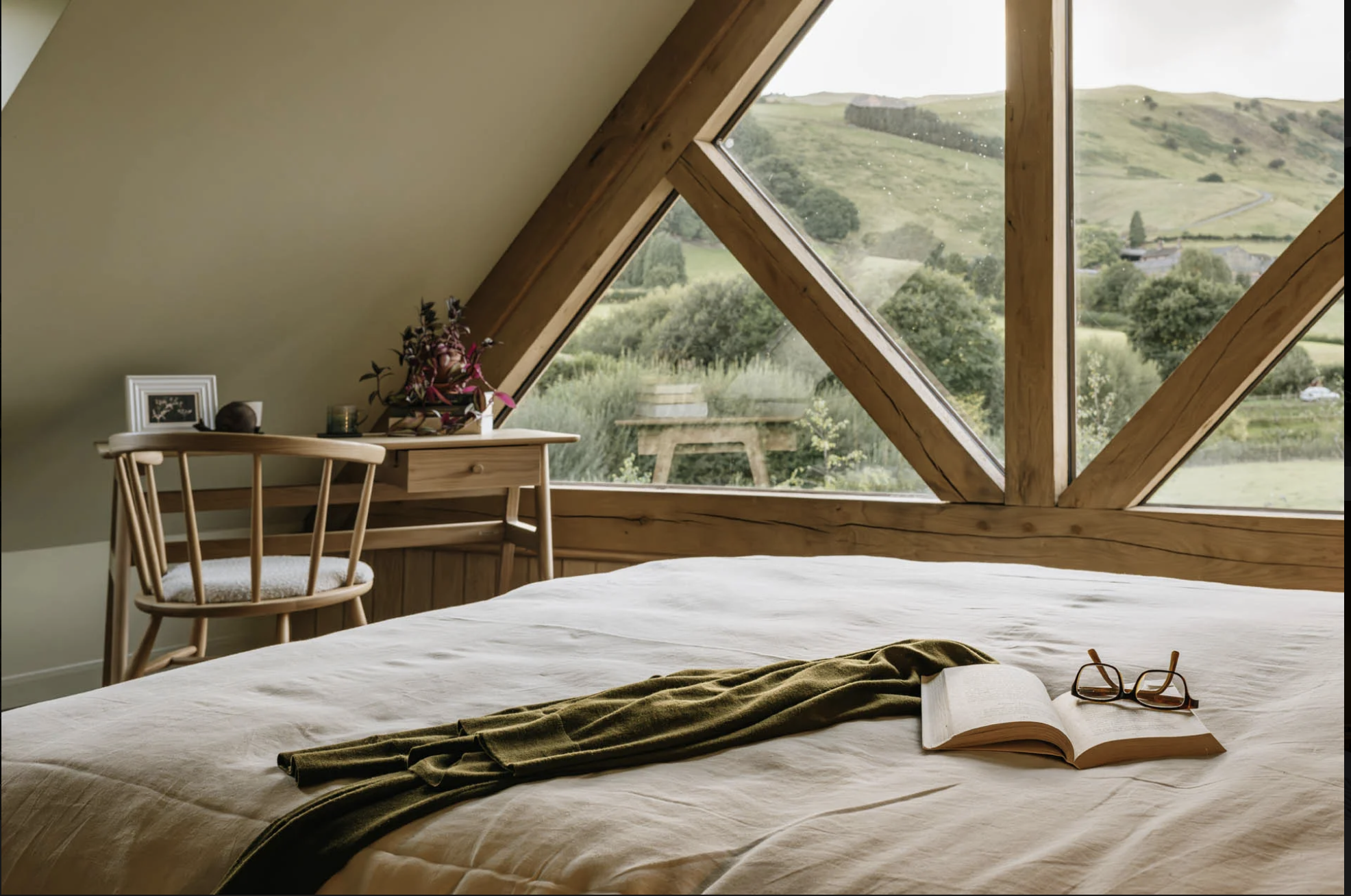 A cozy bedroom with a sloped ceiling and large wooden-framed windows offering a view of green rolling hills and countryside. There is a bed with a white coverlet, an open book, and a pair of glasses on it. A wooden chair and a small desk with decorative items and a flower arrangement are in the corner.