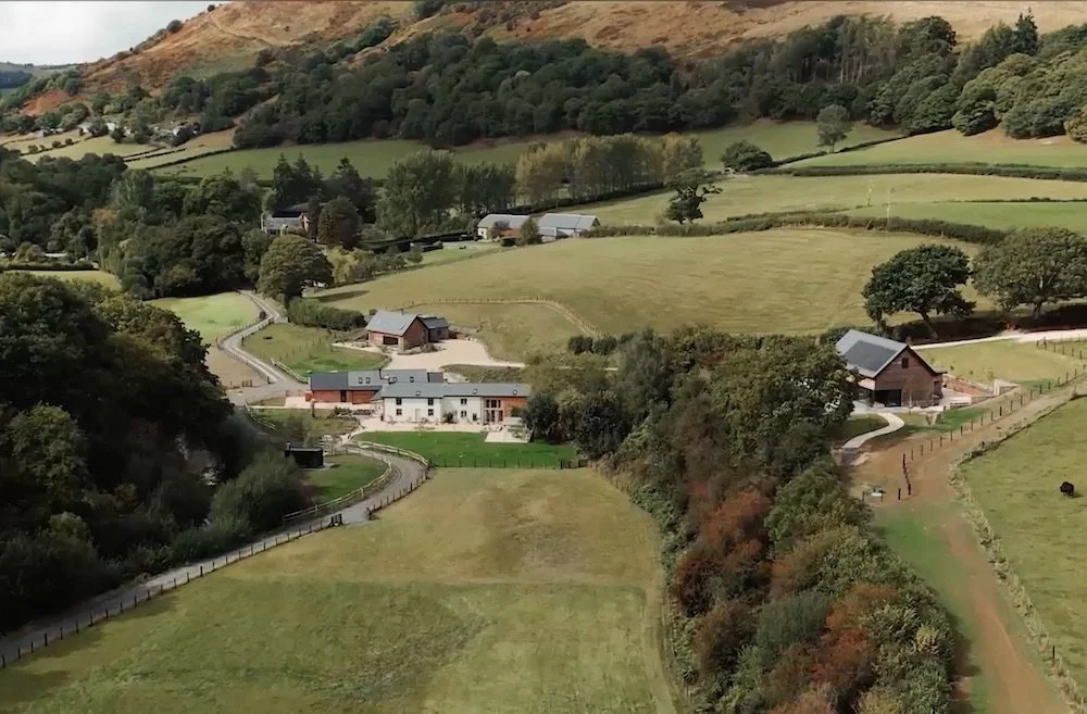Scenic aerial view of countryside with rolling green fields, farm buildings, trees, and distant hills.