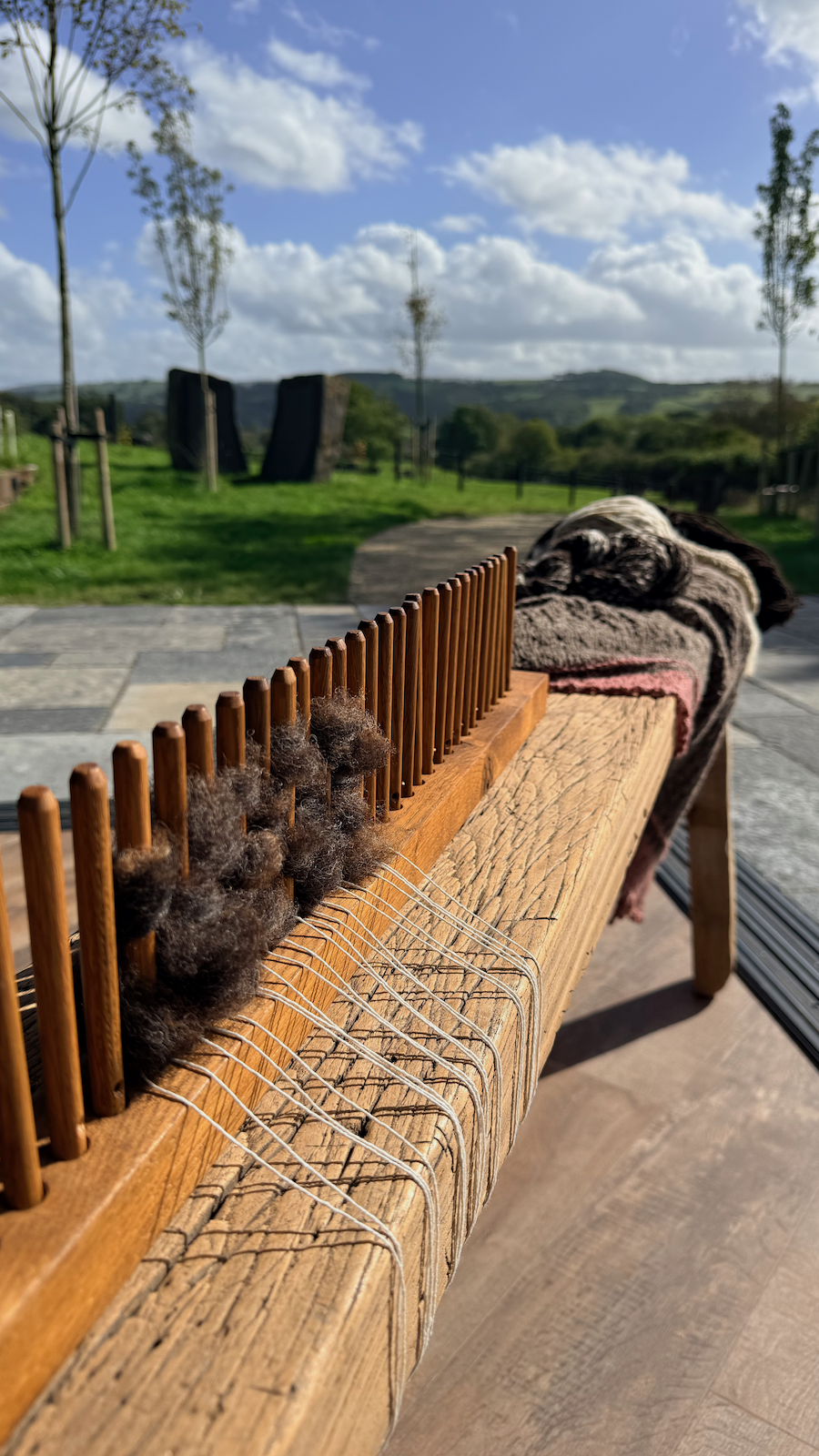 Close-up of a wooden weaving loom with wool fibers, outdoors on a sunny day with a grassy landscape and blue sky in the background.