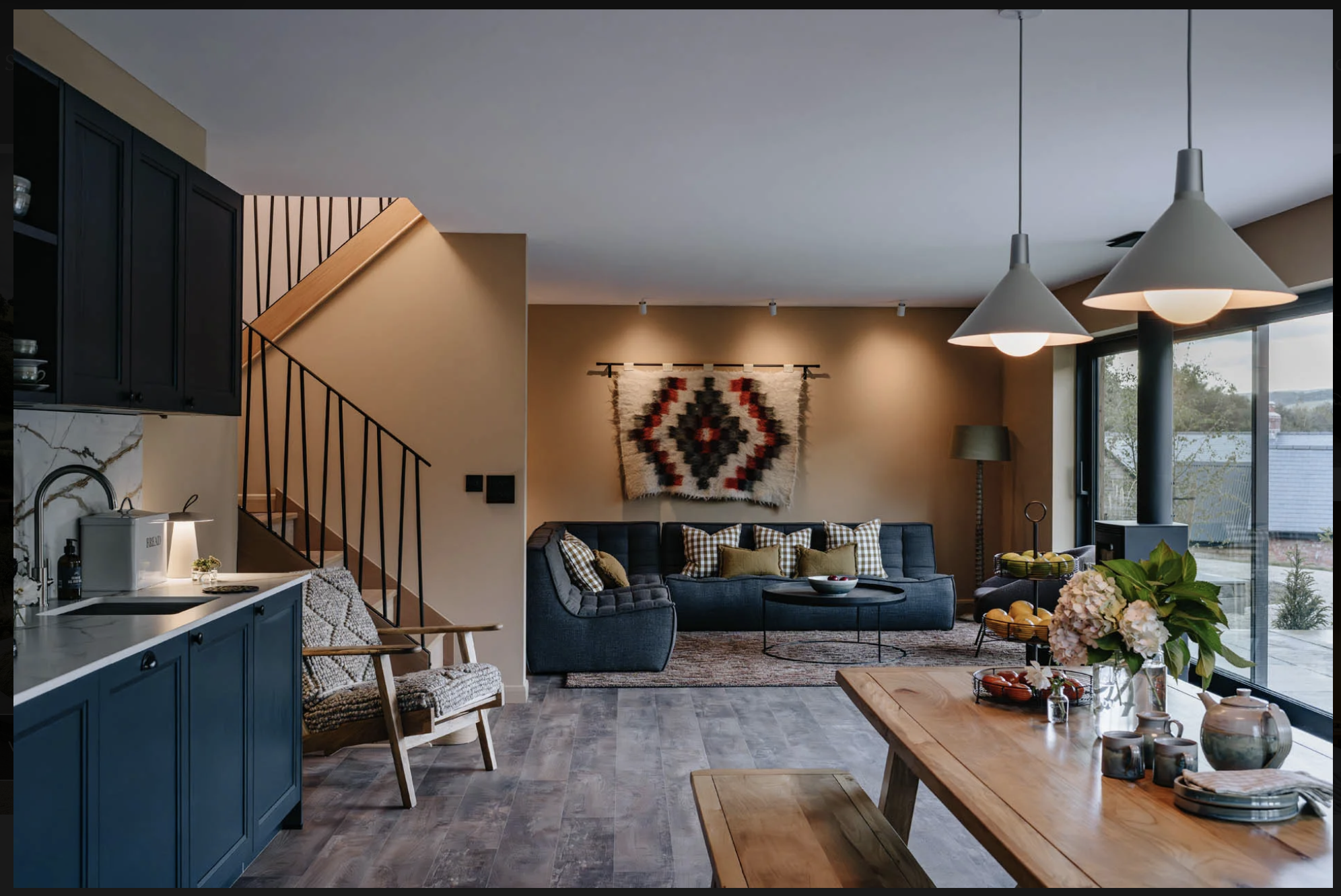 Modern open-concept living room with a navy blue couch, patterned pillows, wall art, large windows, pendant lights, and a wooden dining table with vases and flowers.