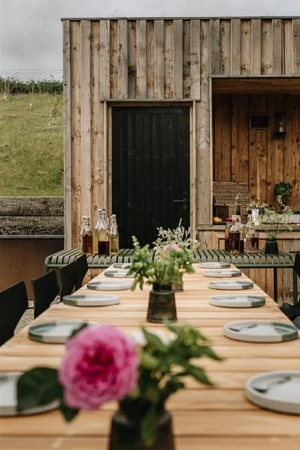 Outdoor dining table set with white plates and a flower arrangement in the foreground, against a wooden building with a black door and bottles of drinks on the table.