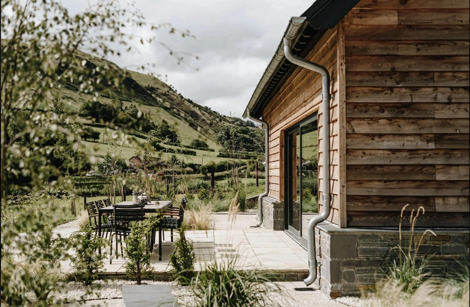 A wooden house with black trim situated in a mountainous area. There is an outdoor patio with a table and chairs, surrounded by greenery and plants. The background features rolling green hills and a cloudy sky.