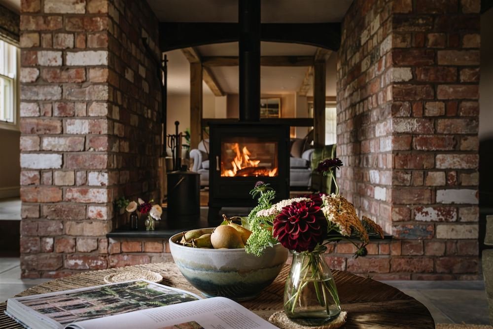 Living room with a brick fireplace and a wood stove burning, decorated with flowers in vases on a wooden table and an open book.