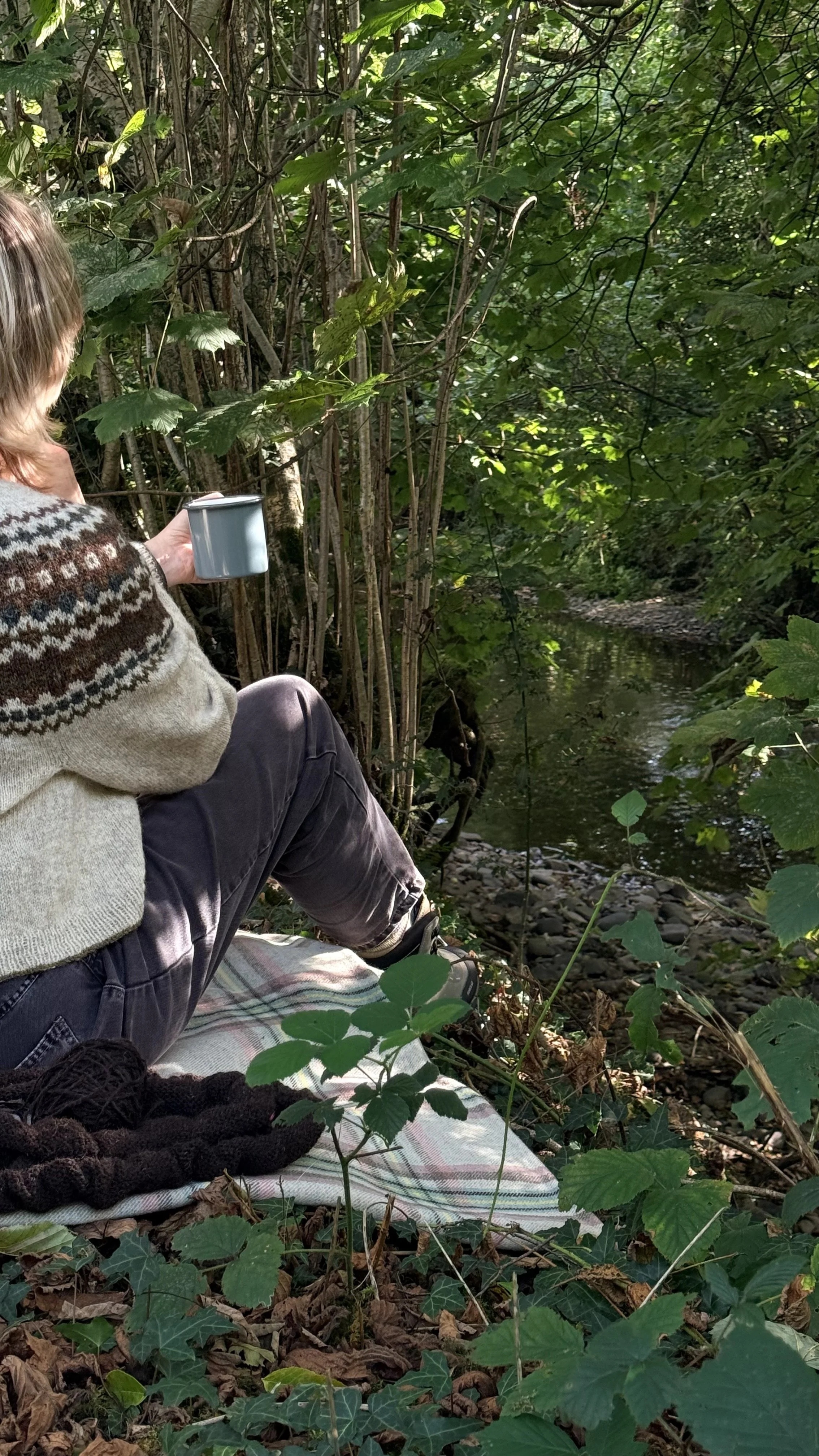 Person sitting outdoors near a stream, surrounded by green foliage, holding a cup, with a blanket and shoes nearby.
