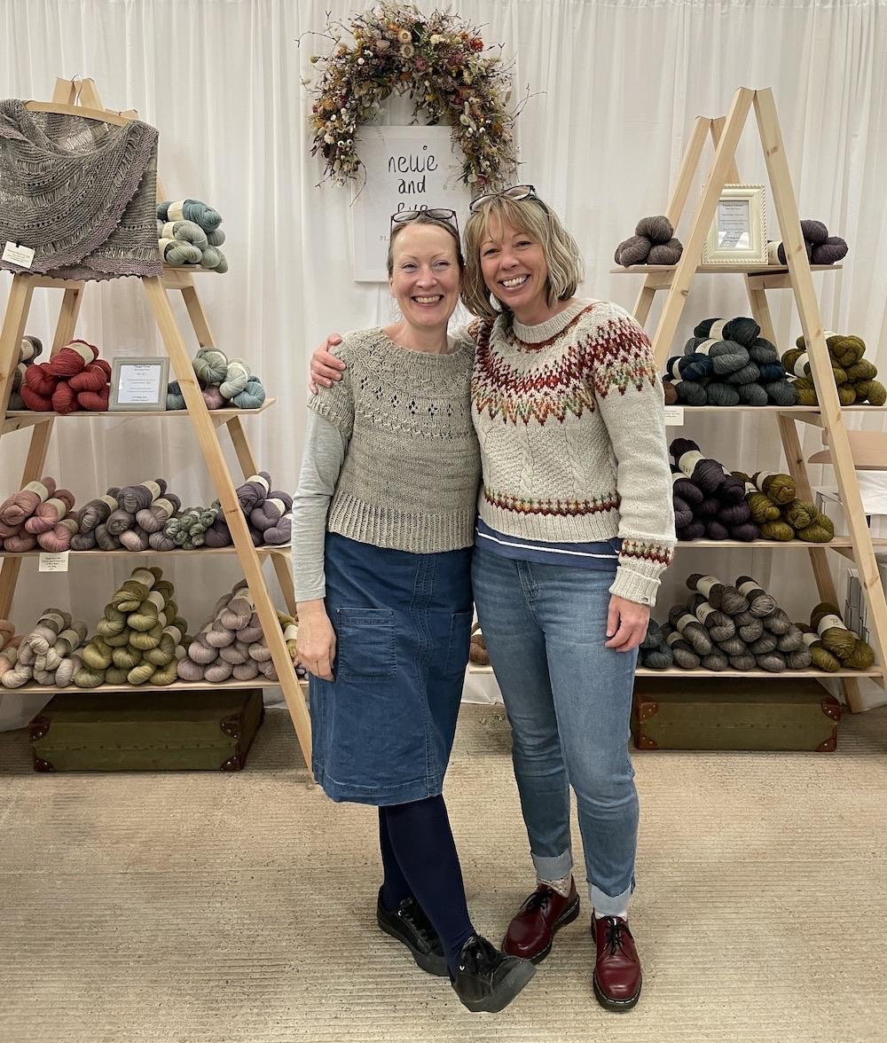 Two women smiling and standing close together with arms around each other in front of shelves filled with yarn balls, in a yarn shop or craft fair, with a white backdrop and decorative wreath above a sign that says ‘Nellie and Eve'