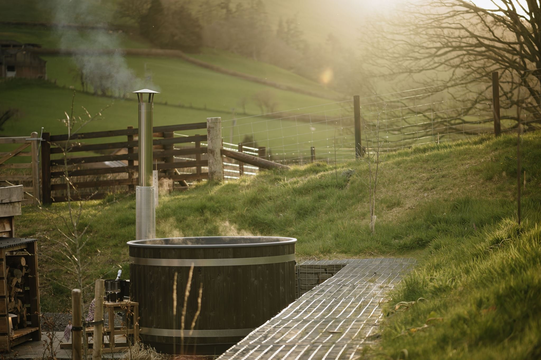 A rural backyard with a fenced area, a metal fire pit, and a grassy hillside. Sunlight filters through trees, creating a warm glow and soft focus in the background.