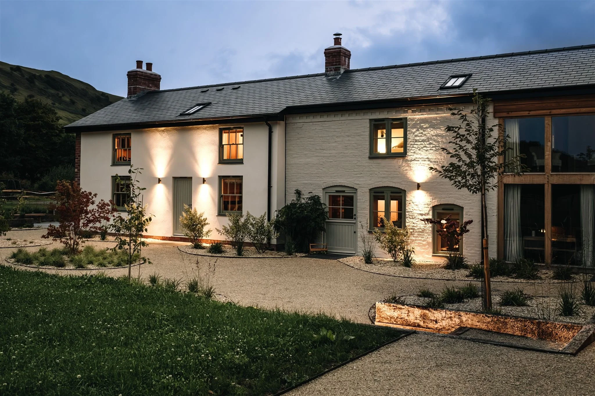 A two-story house with a gray shingled roof and white brick exterior, illuminated by outdoor lighting at dusk. The house has multiple windows with warm interior lighting and a surrounding landscaped garden with small trees and plants. Hills are visible in the background.
