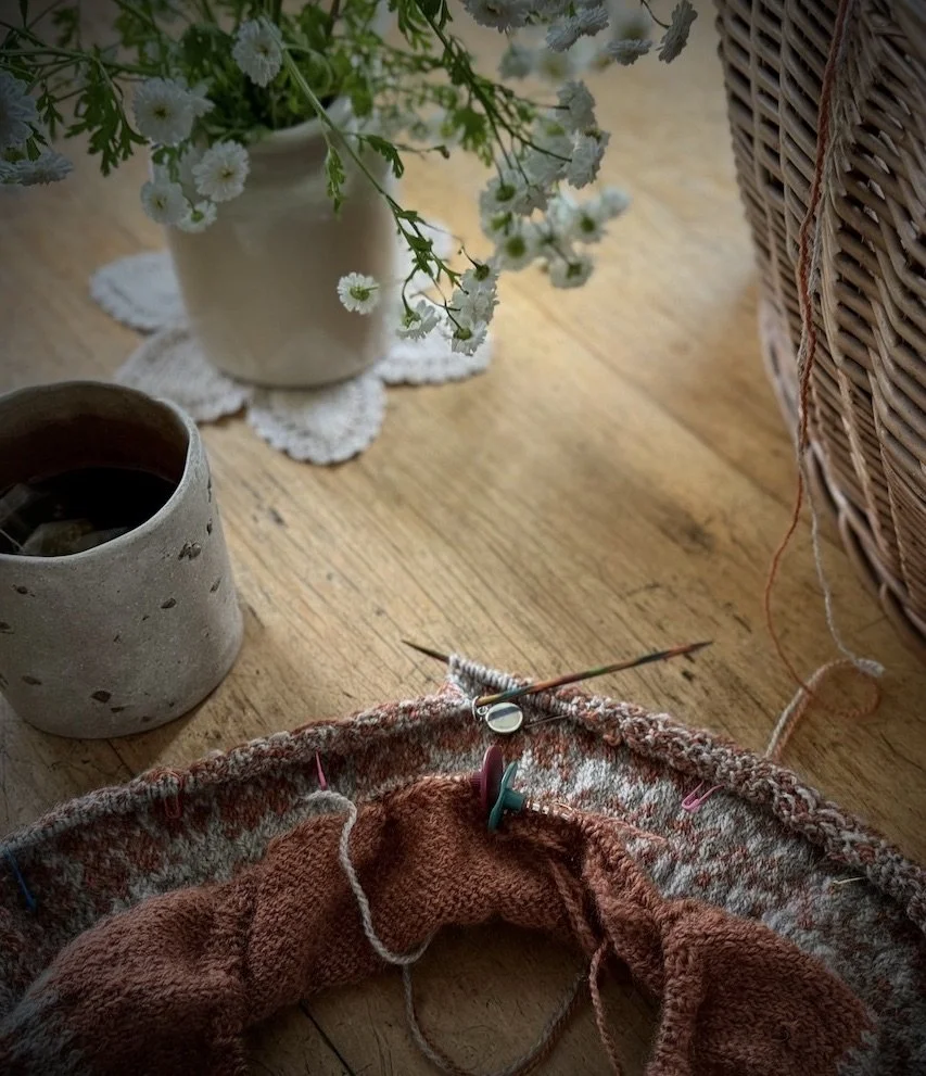 A knitting project in progress with a partially knitted reddish-brown garment on circular knitting needles, placed on a wooden surface. Nearby are a cup of black coffee, a potted white flower, and a wicker basket.