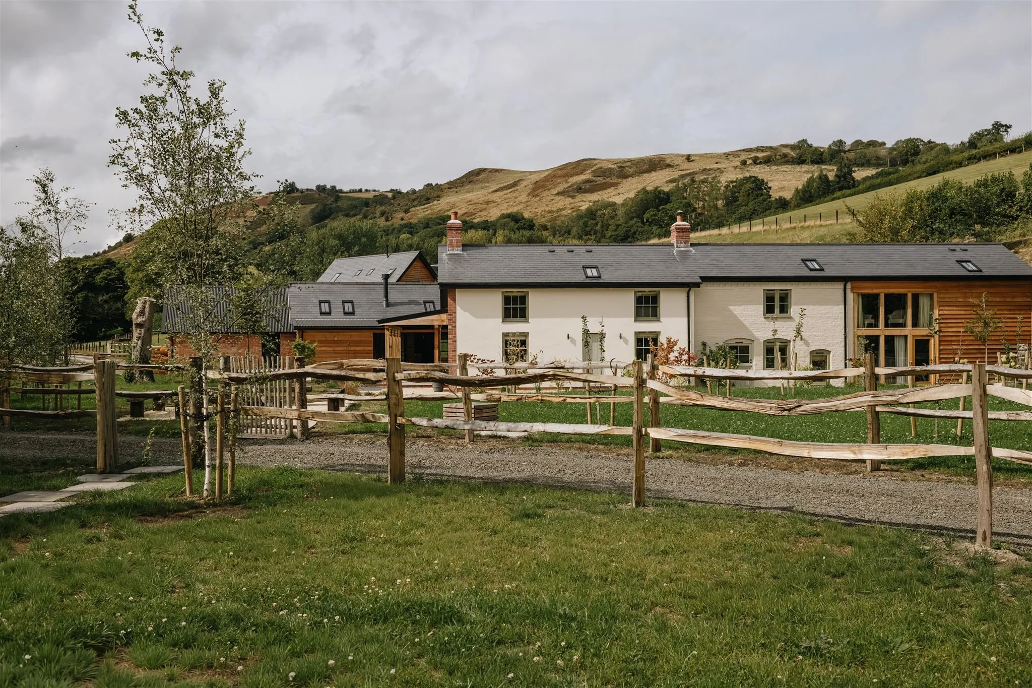 A rural scene featuring a fenced yard with green grass, a few young trees, and a white house with a gray roof and chimney, set against rolling hills under cloudy skies.