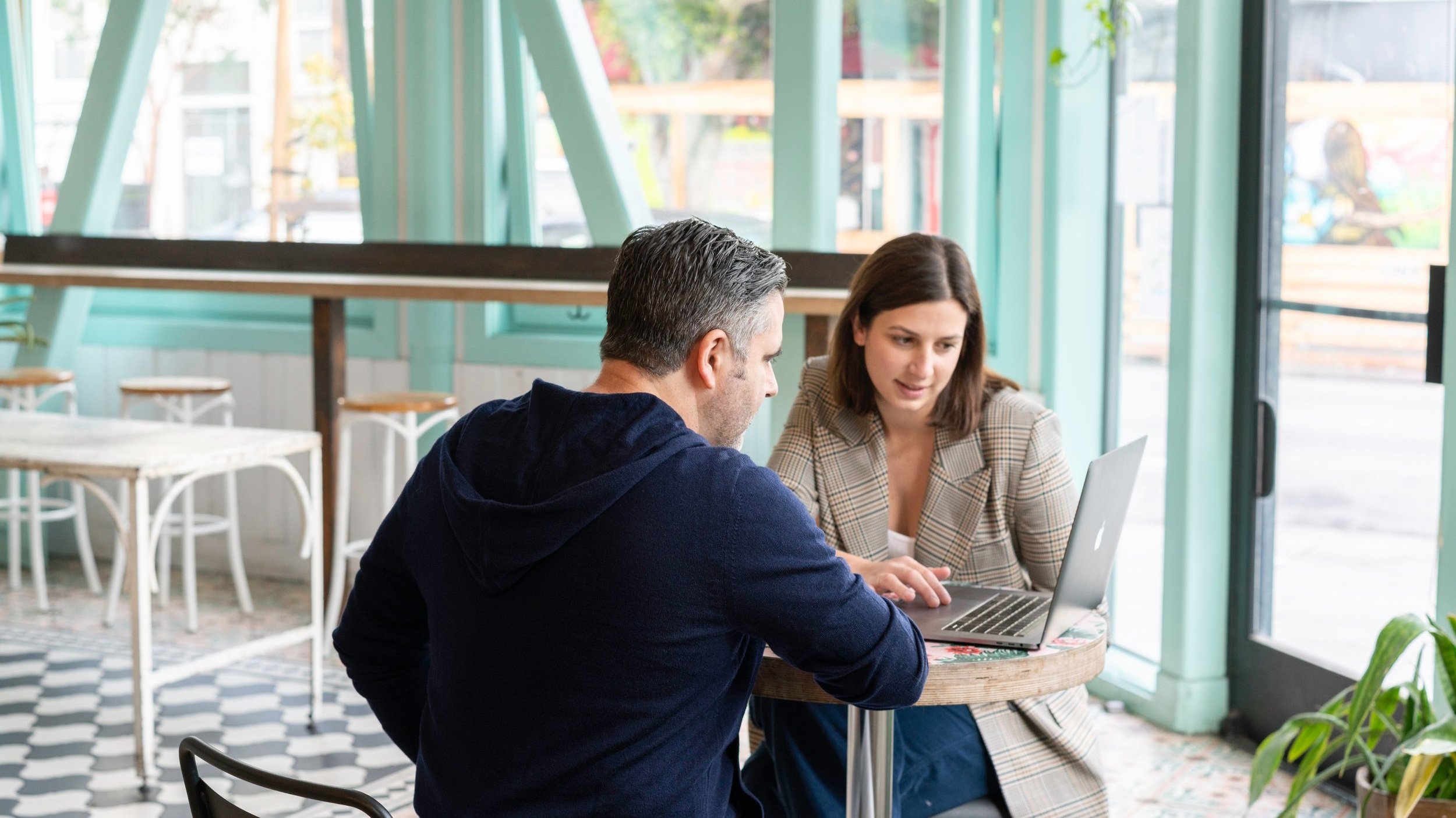 A man and woman sitting at a small round table in a cafe, looking at a laptop together.