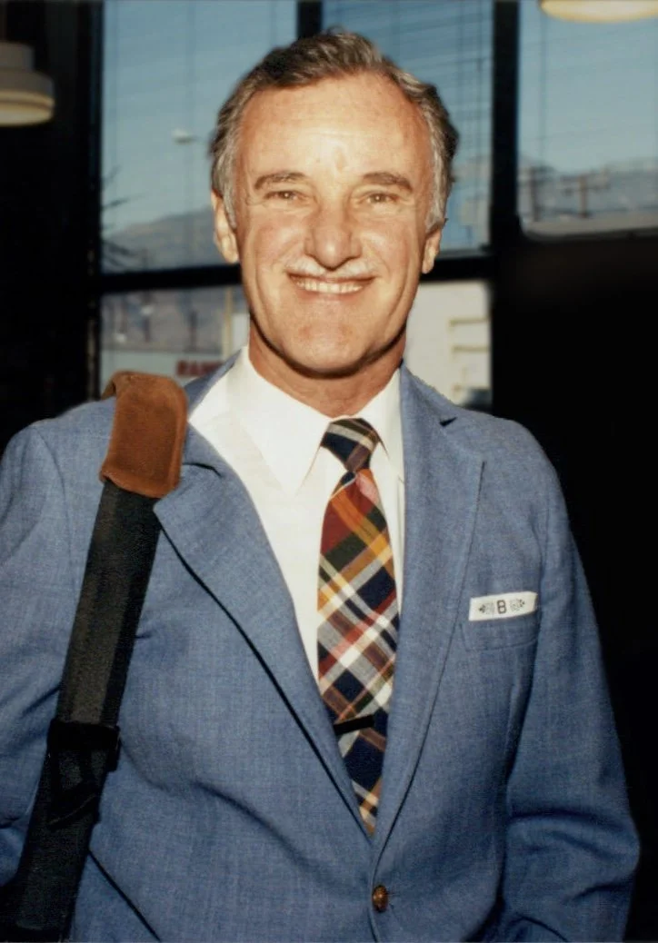 Author and adventurer Bruce Brander in a blue blazer, white shirt, and plaid tie standing indoors with a window behind him, carrying a shoulder bag.