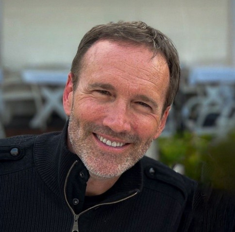 A smiling middle-aged man with short brown hair and a beard, wearing a black jacket, outdoors with blurred background.