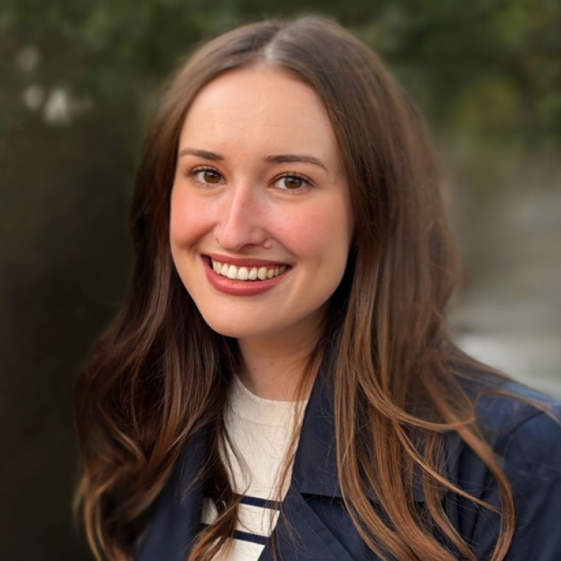 A young woman with long brown hair, smiling outdoors with blurred greenery in the background.