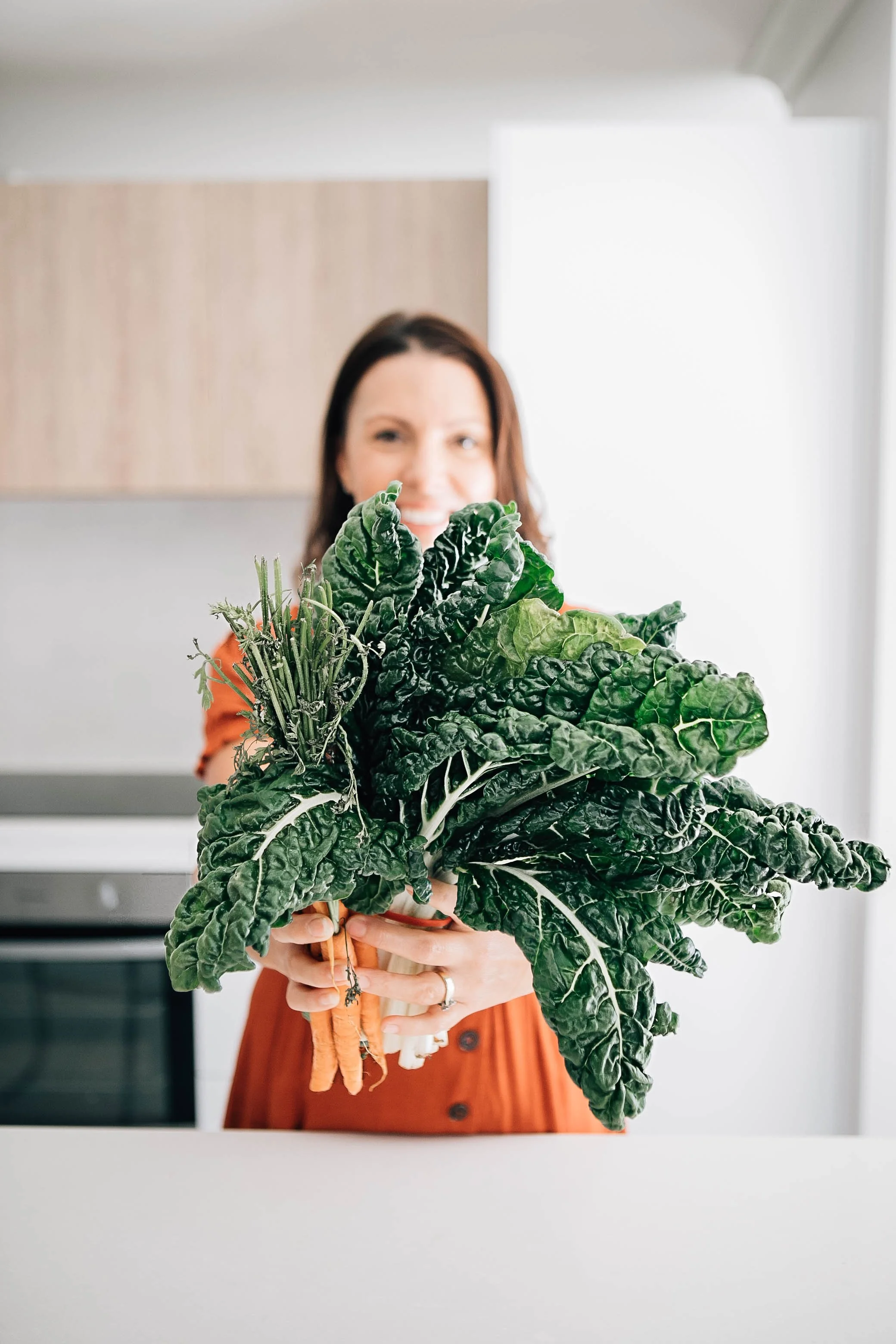 Woman holding a large bunch of dark green leafy vegetables, with a kitchen background.