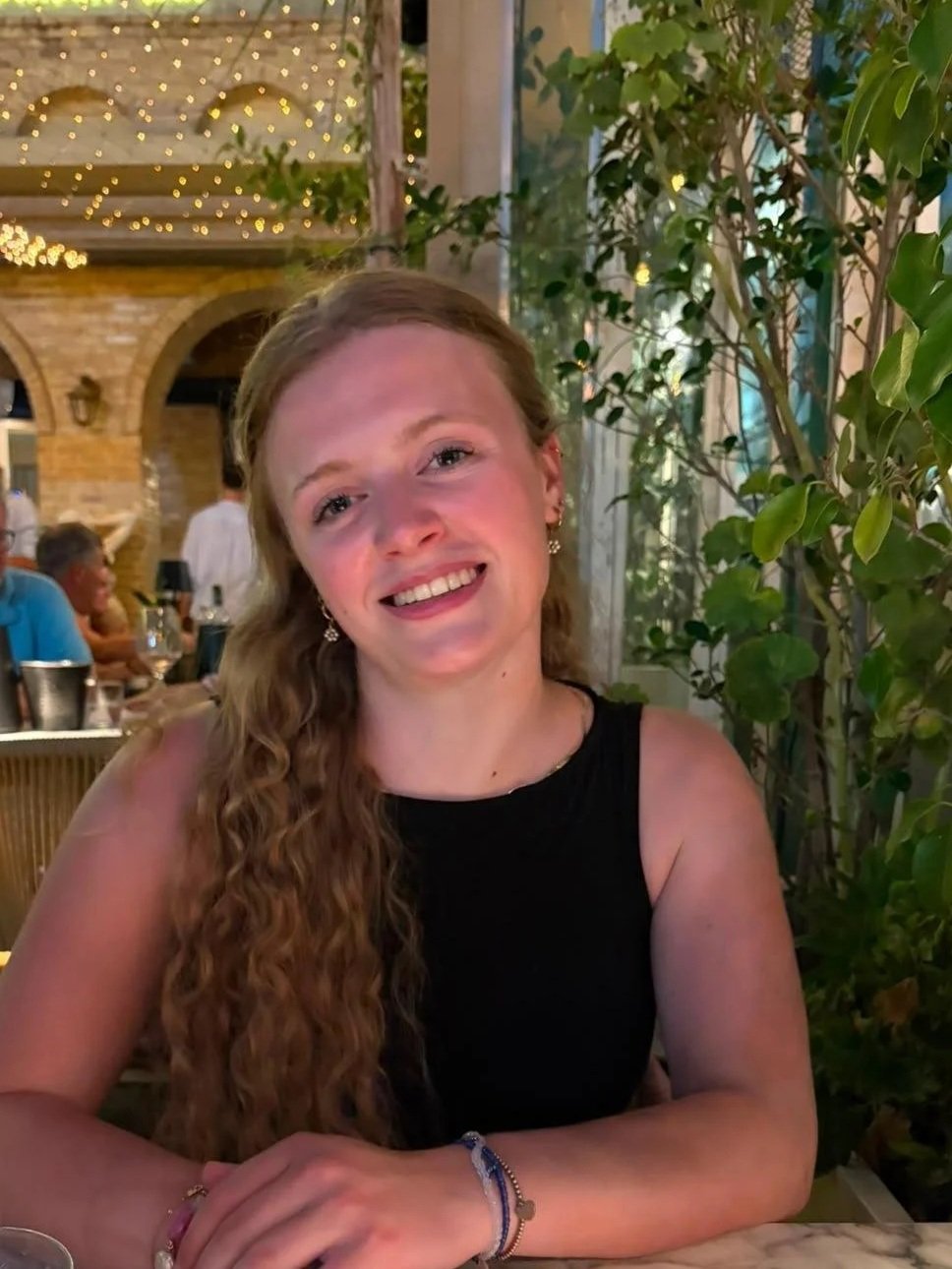Een jonge vrouw met lang, krullend haar glimlacht in een restaurant met décor en planten op de achtergrond.